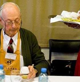 Elderly man wearing glasses and an apron, sitting at a table with a notepad and a bottle of water. - Olive Oil Times