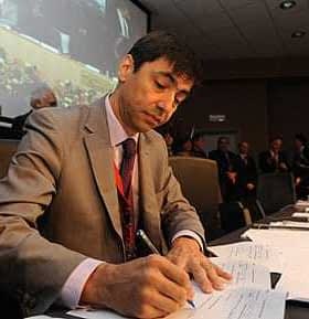 A man in a suit signing documents at a conference table with others in the background. - Olive Oil Times