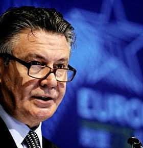 Karel De Gucht delivering a speech at a podium during a conference with a blue background. - Olive Oil Times