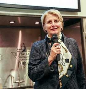 Woman holding a microphone and speaking at a culinary event with a scarf around her neck. - Olive Oil Times