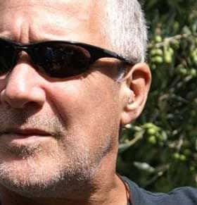 Homme aux cheveux gris courts portant des lunettes de soleil, avec des oliviers en arrière-plan. - Olive Oil Times