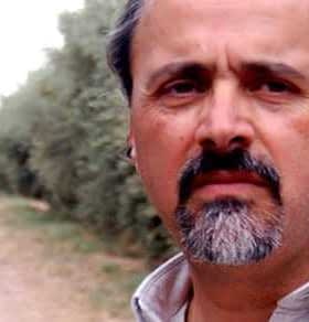 A man standing in an olive grove with trees lining a dirt path in the background. - Olive Oil Times