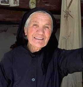 Elderly woman wearing a black dress and headscarf, smiling while leaning against a wall. - Olive Oil Times