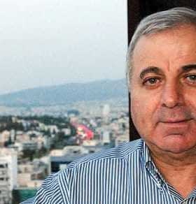 A man with gray hair wearing a striped shirt, posing for a portrait with a cityscape in the background. - Olive Oil Times
