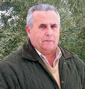 Un homme portant une veste verte et un pull beige debout à l'extérieur devant de la verdure. - Olive Oil Times