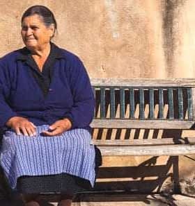 Older woman wearing a blue sweater and skirt sitting on a wooden bench against a wall. - Olive Oil Times