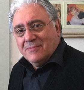 Un homme aux cheveux gris et aux lunettes portant une veste noire, debout devant une œuvre d'art encadrée. - Olive Oil Times