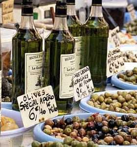 Several bottles of olive oil displayed at a market stall with various types of olives in bowls. - Olive Oil Times