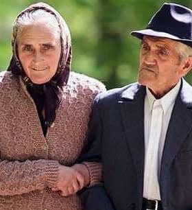 Elderly couple holding hands while walking outdoors, dressed in traditional clothing. - Olive Oil Times