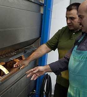 Two men inspecting a baking oven with bread inside, one wearing an apron and the other in a green shirt. - Olive Oil Times