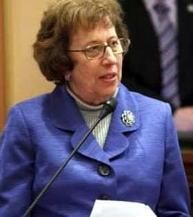 Eine Frau mit lockigem Haar, Brille und blauem Anzug spricht an einem Podium. - Olive Oil Times