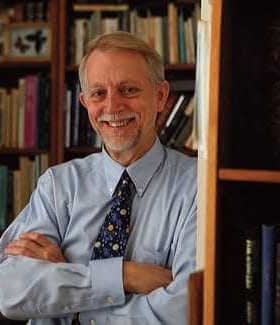 A man with gray hair and glasses smiling while standing in front of bookshelves filled with books. - Olive Oil Times
