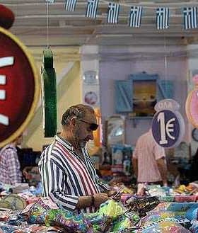 A market scene featuring price tags displaying '1€' and shoppers browsing various items. - Olive Oil Times