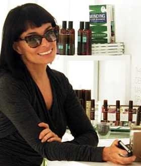 Woman with dark hair and sunglasses smiling while leaning against a counter with products in the background. - Olive Oil Times