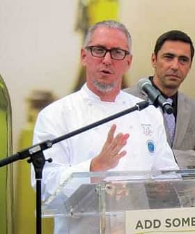 A chef in a white coat speaking at a podium with a microphone during an event. - Olive Oil Times
