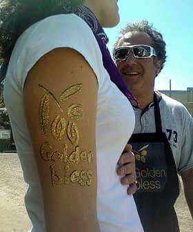 A person's arm with golden body art displaying the text 'Golden Bless'. - Olive Oil Times