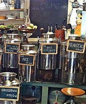 Various stainless steel containers for olive oil displayed on a shelf with labels indicating different types. - Olive Oil Times