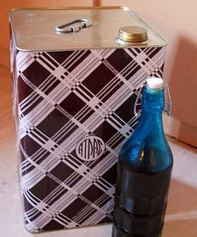 A square metal container with a patterned design next to a blue glass bottle. - Olive Oil Times