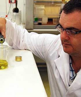 Scientist in a lab coat pouring olive oil from a bottle into a beaker in a laboratory setting. - Olive Oil Times