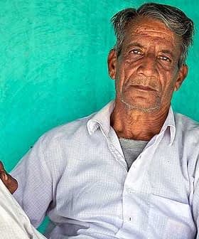 An elderly Indian farmer sitting against a turquoise wall, wearing a white shirt and pants. - Olive Oil Times