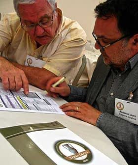 Two men discussing and reviewing documents at a table, one pointing at the papers. - Olive Oil Times