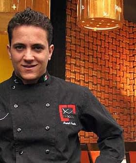 Chef wearing a black culinary uniform with a logo, standing in a restaurant setting. - Olive Oil Times