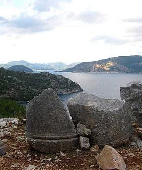 Three large stone fragments positioned near the coastline with a view of the sea and mountains in the background. - Olive Oil Times
