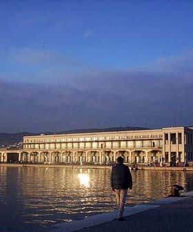 Trieste Maritime Station building reflecting on the water during sunset with a person walking nearby. - Olive Oil Times