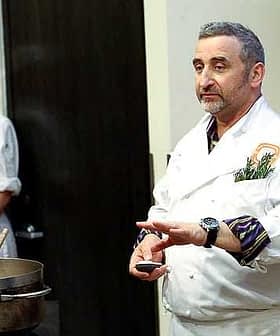 Chef in a white coat demonstrating cooking techniques with a pot in front of him and a female chef in the background. - Olive Oil Times