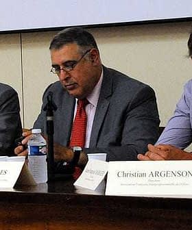 Three men seated at a conference table with water bottles and nameplates in front of them. - Olive Oil Times