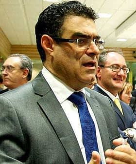 A man in a gray suit with a blue tie speaking in a group setting with other men in suits. - Olive Oil Times