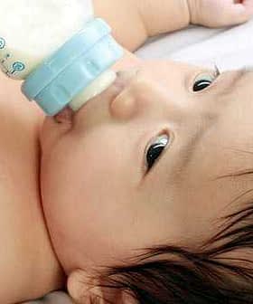 Infant with dark hair drinking from a blue baby bottle while lying down. - Olive Oil Times