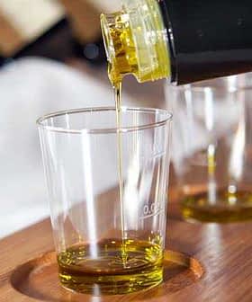 Olive oil being poured from a bottle into a clear glass sample cup on a wooden tray. - Olive Oil Times