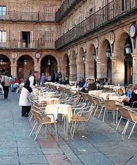 Outdoor dining area with tables and chairs set up in a plaza, featuring people seated and a server attending to guests. - Olive Oil Times