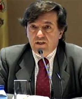 Man in a suit speaking at a conference with a Spanish flag visible in the background. - Olive Oil Times