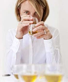 Woman holding a glass of olive oil while tasting it, with several glasses in front of her. - Olive Oil Times