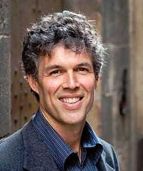 A man with curly hair wearing a dark suit and smiling at the camera. - Olive Oil Times