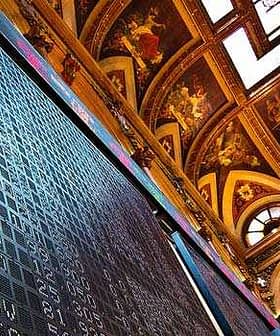 Large electronic stock market display board in a historic building with ornate ceiling. - Olive Oil Times