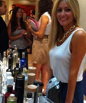 Woman with long blonde hair standing at a table filled with various product bottles at an event. - Olive Oil Times