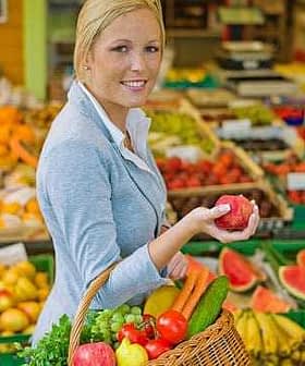 Woman in a gray blazer holding a red apple while carrying a basket of fresh fruits and vegetables. - Olive Oil Times