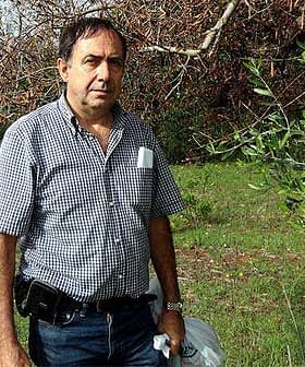 Man wearing a checkered shirt and holding bags while standing outdoors in a green area. - Olive Oil Times