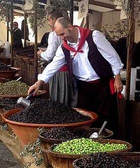 A man in traditional attire selecting olives from large bowls at a market stall. - Olive Oil Times