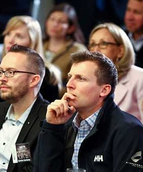 Two men in the foreground listening attentively during an event with an audience in the background. - Olive Oil Times