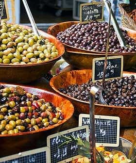 Multiple wooden bowls filled with different types of olives displayed at a market. - Olive Oil Times