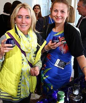 Two women holding glasses at a social event, one wearing a yellow vest and the other in a floral dress. - Olive Oil Times