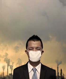Businessman in a suit and mask standing against an industrial backdrop with smoke stacks and cloudy sky. - Olive Oil Times