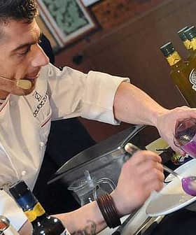 Chef in a white coat preparing a dish with olive oil and colorful ingredients on a countertop. - Olive Oil Times