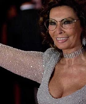 Woman wearing a sparkling silver dress and glasses, smiling and waving at an event. - Olive Oil Times