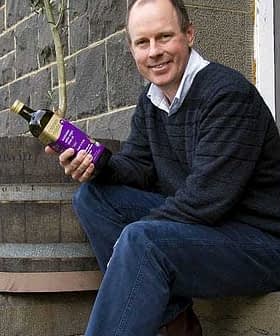 Man sitting on a step holding a bottle of olive oil with a tree in the background. - Olive Oil Times