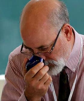 A man with glasses and a beard smelling a blue container in a classroom setting. - Olive Oil Times